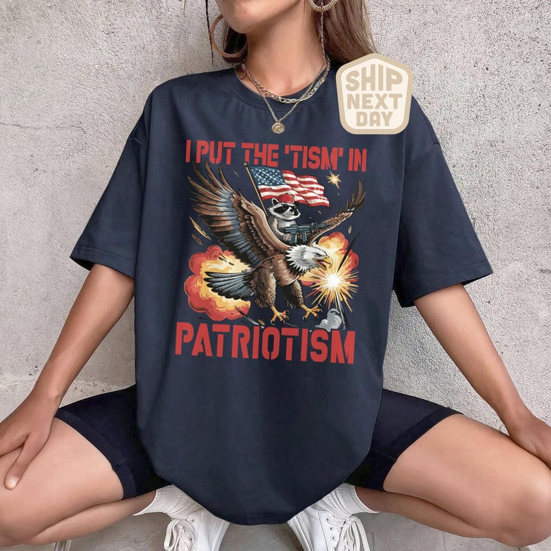 Puede incluir: Camiseta azul marino con un dise&ntilde;o patri&oacute;tico. Muestra un &aacute;guila llevando un mapache con una bandera estadounidense y el texto "I PUT THE 'TISM' IN PATRIOTISM". El dise&ntilde;o incluye explosiones y chispas.