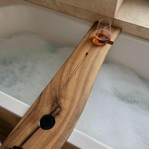 May include: A wooden bathtub tray with a glass of amber liquid resting on it. The tray has a dark, natural wood grain finish and a circular cutout. A black washcloth is placed on the tray. The tray is positioned over a bathtub filled with bubbles.