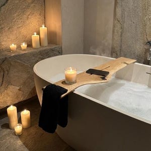 Puede incluir: Una escena de baño serena con una bañera blanca llena de burbujas. Una bandeja de baño de madera sostiene una vela encendida y un objeto negro. Una toalla negra cuelga de la bandeja. Varias velas encendidas de diferentes tamaños están dispuestas en un estante de piedra y en el suelo.