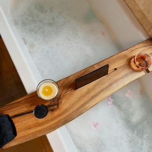 Puede incluir: Una bandeja de bañera de madera con una vela, una taza de té y un soporte para teléfono negro. La bandeja se coloca sobre una bañera blanca llena de burbujas y pétalos de flores rosas. El baño tiene baldosas beige.