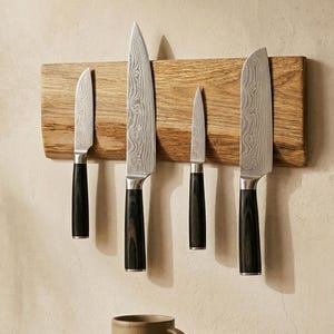 May include: Five kitchen knives with patterned blades and dark wooden handles are displayed on a wooden magnetic strip. The knives are of different sizes and shapes. A neutral-toned mug is visible below the knives.