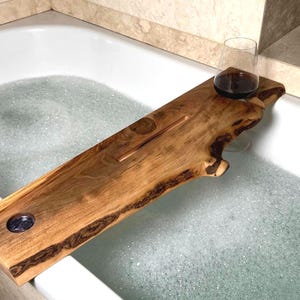 Puede incluir: Una bandeja de baño de madera con borde natural, diseñada para sostener una copa de vino. La bandeja, hecha de madera de tonos cálidos, tiene una incrustación de piedra oscura y se apoya sobre una bañera blanca llena de burbujas. La longitud de la bandeja es de unos 76 cm.