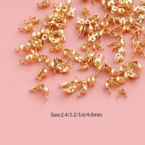 May include: A collection of gold-colored metal crimp covers used for jewelry making. These small, hollow, hemispherical findings feature a loop at the top for attachment. The text printed at the bottom indicates the sizes: 2.4/3.2/3.6/4.0mm.