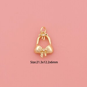 May include: A gold-toned metal charm shaped like a bikini top with a small bow detail and a clear stone accent. The charm includes a jump ring at the top for attachment. The text printed on the image indicates a size of 21.3x12.2x6mm.