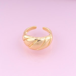 Puede incluir: Un anillo dorado con una banda gruesa y texturizada. La banda tiene un diseño ondulado y acanalado.