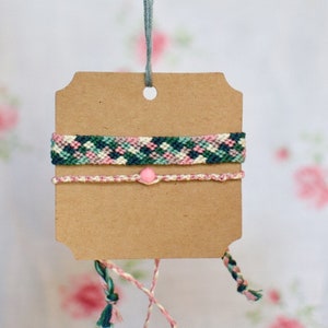 May include: Two friendship bracelets with a pink bead. The top bracelet is green, pink, and white. The bottom bracelet is pink and white. The bracelets are hanging from a brown card with a hole in the top.
