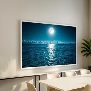 May include: A framed print of a moonlit ocean scene. The image features a bright full moon in a dark blue sky, reflecting a shimmering path of light on the water. White clouds line the horizon, and the water's surface is a deep blue.