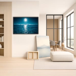 May include: A large framed print of a moonlit ocean scene with a bright moon reflecting on the water. The room features a neutral color palette with a white ottoman, a canvas print, and a wooden frame. A dining table and chairs are visible in the background.
