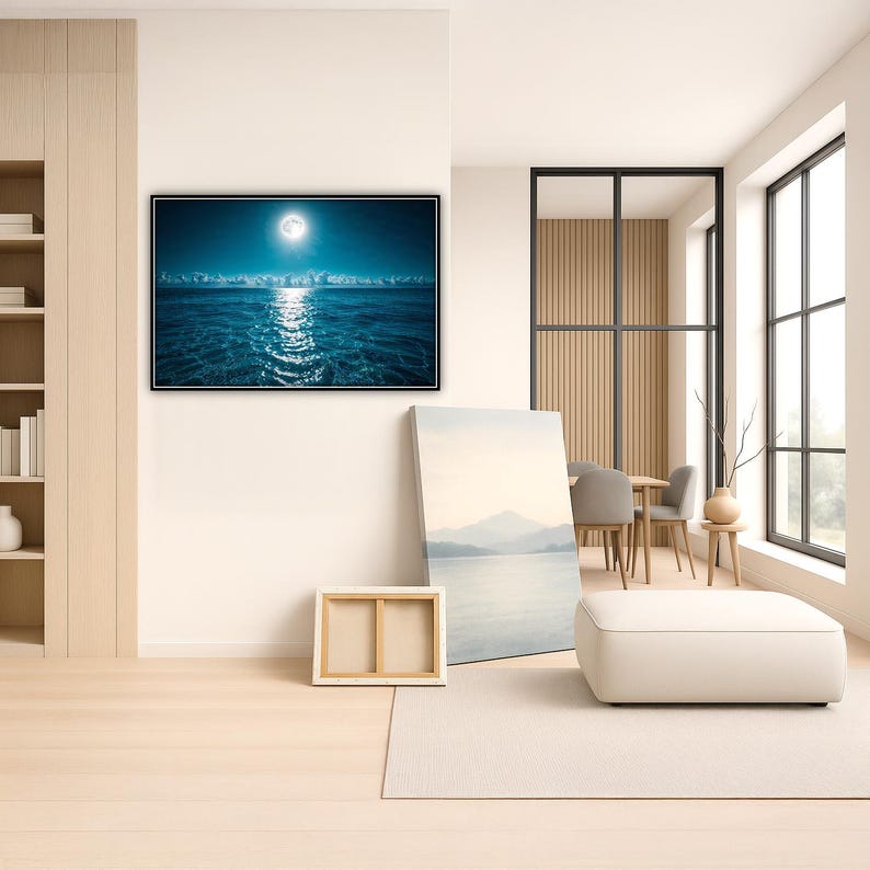 May include: A large framed print of a moonlit ocean scene with a bright moon reflecting on the water. The room features a neutral color palette with a white ottoman, a canvas print, and a wooden frame. A dining table and chairs are visible in the background.