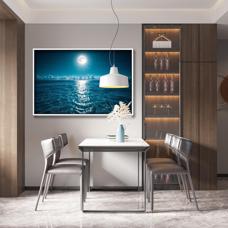 May include: A dining room scene featuring a framed seascape painting with a bright moon reflecting on the water. A white table and two gray chairs are in front of the painting. A white pendant light hangs above the table. A cabinet with wine bottles is on the right.