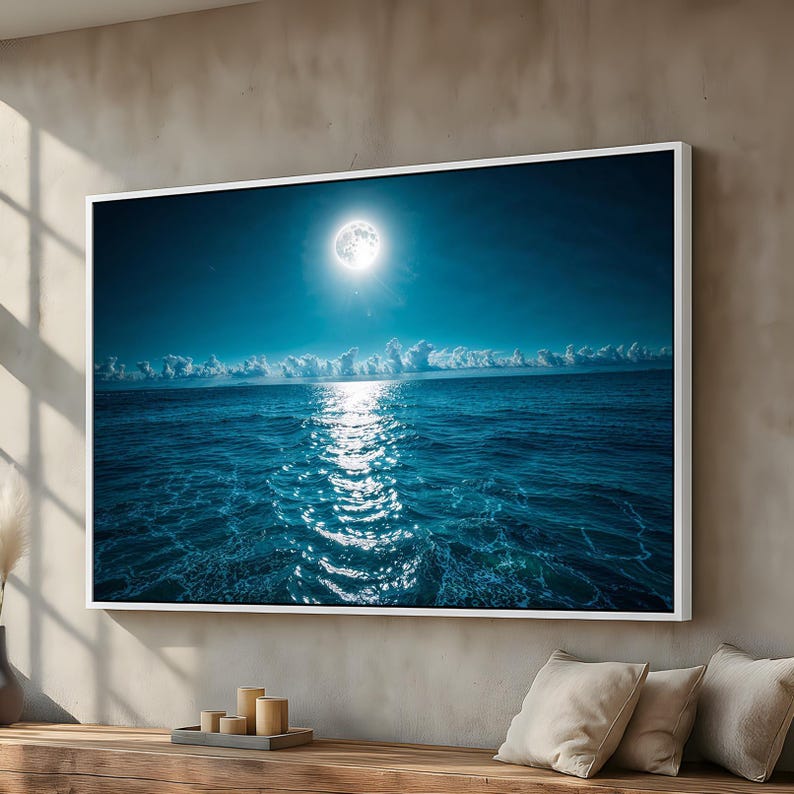 May include: A framed print depicts a nighttime ocean scene. A bright full moon illuminates the dark blue sky and reflects on the water. White clouds line the horizon. The print is in a white frame.