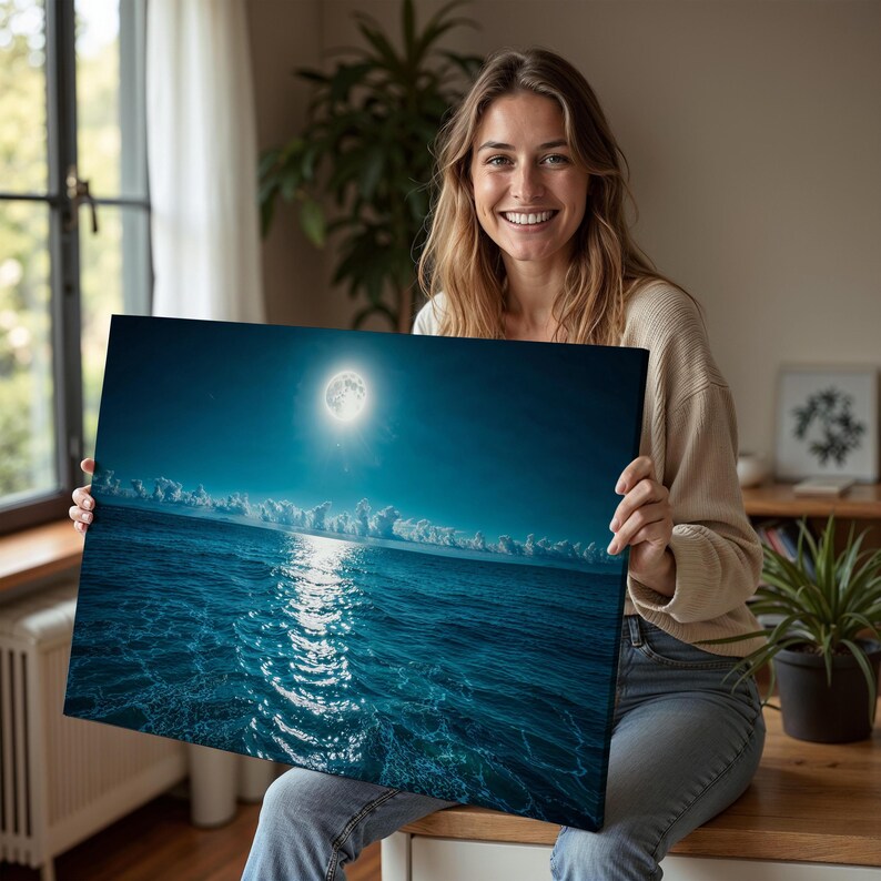 May include: A canvas print depicting a nighttime ocean scene under a full moon. The artwork features a bright moon in a dark blue sky, with the moon's reflection shimmering on the water's surface. The print is held by a person.