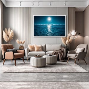 May include: A modern living room with a large framed artwork depicting a moonlit ocean scene. The room features a beige sofa, two armchairs, and two round ottomans. A neutral-toned rug and decorative pampas grass complete the decor.