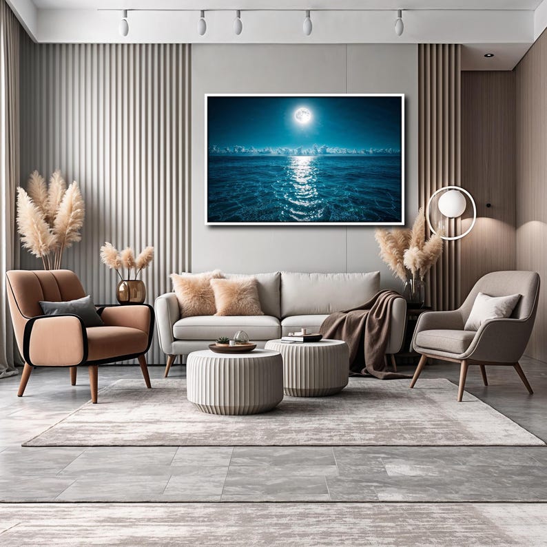 May include: A modern living room with a large framed artwork depicting a moonlit ocean scene. The room features a beige sofa, two armchairs, and two round ottomans. A neutral-toned rug and decorative pampas grass complete the decor.