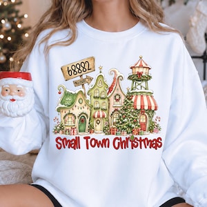 May include: A white long-sleeve sweatshirt featuring a graphic of a small town with Christmas decorations, a sign reading 68882 Nashville TN, and the text Small Town Christmas in red. A Santa Claus mug is held in the foreground of the image.