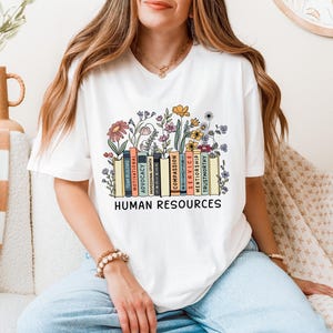Op de afbeelding: Wit T-shirt met een afbeelding van een stapel boeken met bloemen die eruit groeien. De boeken hebben woorden op hun ruggen, waaronder "compassion", "service", "mentorship" en "trustworthy". De tekst "HUMAN RESOURCES" is onder de boeken gedrukt.