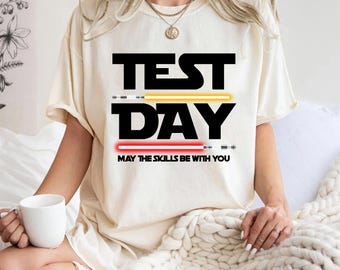 Könnte beinhalten: Ein weißes T-Shirt mit schwarzem Text, der "TEST DAY" lautet, mit einer roten und gelben Lichtschwertgrafik und dem Text "MAY THE SKILLS BE WITH YOU".