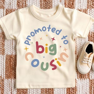 Peut inclure: T-shirt de bébé crème avec l'inscription "promu grand cousin" dans un design circulaire coloré. La chemise est associée à une paire de baskets en toile beige, le tout sur un tapis à motifs.