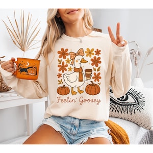 Peut inclure: T-shirt crème à manches longues avec un motif d'oie portant une écharpe et tenant une tasse de café, entourée de citrouilles et de fleurs. Le texte "Feelin' Goosey" est en dessous. Une tasse orange avec un motif d'Halloween est tenue dans l'image.