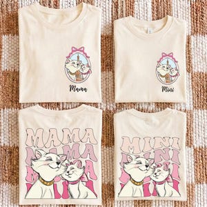 Op de afbeelding: Vier crèmekleurige t-shirts met kattenafbeeldingen. Twee shirts zeggen "Mama" en twee zeggen "Mini". De afbeeldingen bevatten een kat met een roze strik en de woorden "Mama" en "Mini" in een retro lettertype. De shirts zijn opgevouwen en gerangschikt op een patroon.