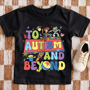 Puede incluir: Camiseta negra con el texto "TO AUTISM AND BEYOND" en letras de colores. La camiseta presenta personajes de dibujos animados y piezas de rompecabezas. Un par de zapatillas de deporte beige est&aacute; al lado de la camiseta.