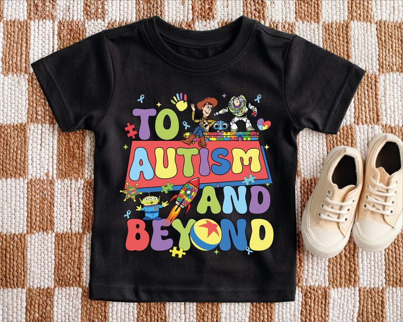 Puede incluir: Camiseta negra con el texto "TO AUTISM AND BEYOND" en letras de colores. La camiseta presenta personajes de dibujos animados y piezas de rompecabezas. Un par de zapatillas de deporte beige est&aacute; al lado de la camiseta.