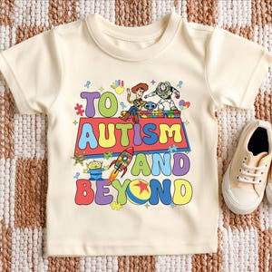 Puede incluir: Camiseta color crema con la frase "TO AUTISM AND BEYOND" en letras coloridas y divertidas. La camiseta presenta personajes de dibujos animados y un gr&aacute;fico de cohete. Un par de zapatillas de lona beige est&aacute;n al lado de la camiseta.