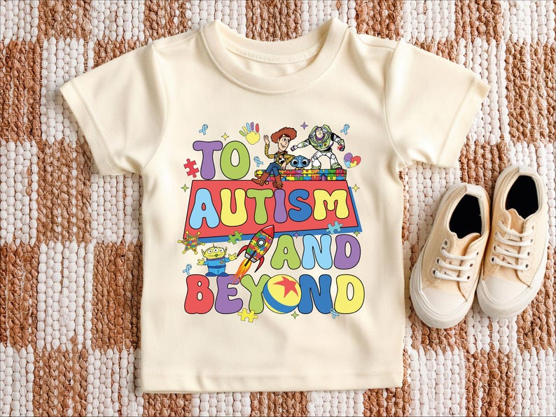 Puede incluir: Camiseta color crema con la frase "TO AUTISM AND BEYOND" en letras coloridas y divertidas. La camiseta presenta personajes de dibujos animados y un gr&aacute;fico de cohete. Un par de zapatillas de lona beige est&aacute;n al lado de la camiseta.