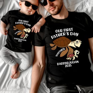 May include: Black t-shirts with a turtle graphic. The shirts read "OUR FIRST FATHER'S DAY TOGETHER DADDY & JULIAN 2025". One shirt is on a baby, the other on an adult.