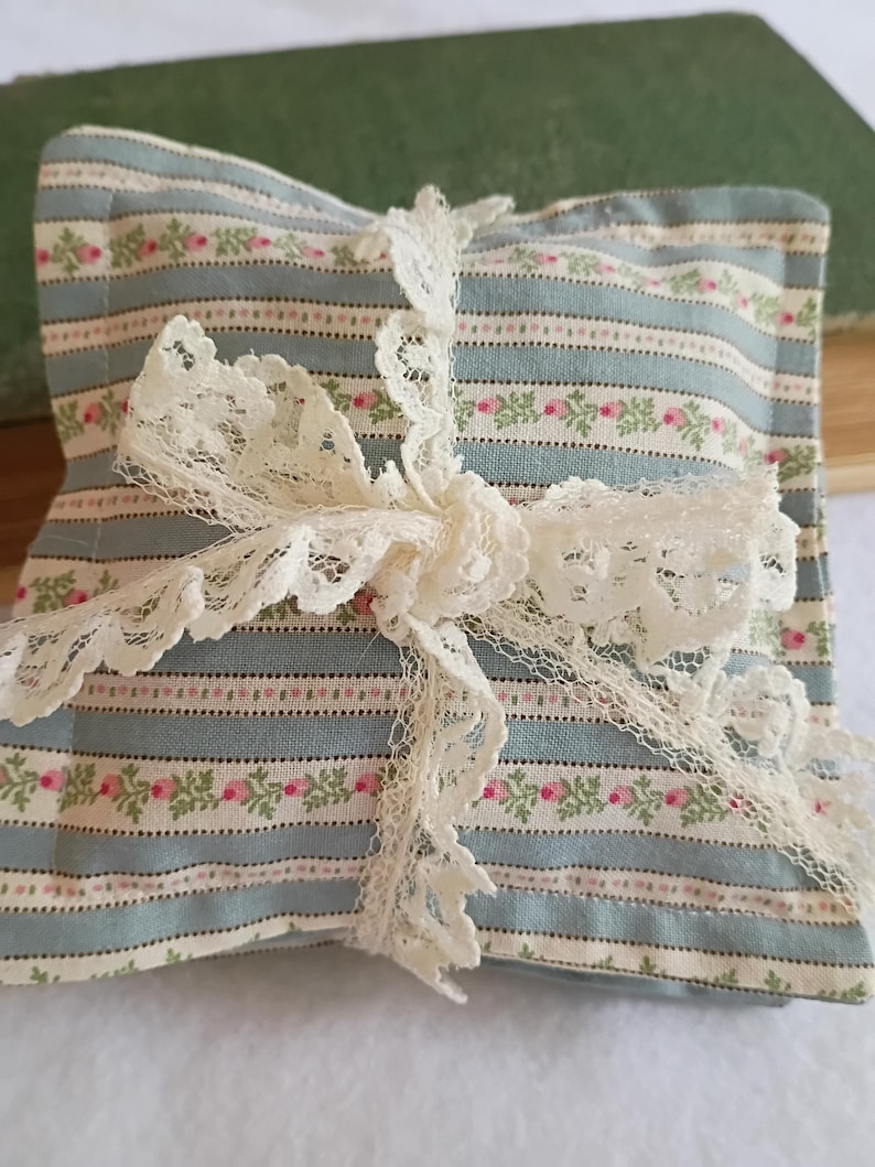 Peut inclure: Un sachet carr&eacute; &agrave; motifs floraux, avec un fond ray&eacute; bleu et blanc, orn&eacute; de roses roses et de feuilles vertes. Le sachet est nou&eacute; avec un ruban de dentelle cr&egrave;me d&eacute;licat, cr&eacute;ant une esth&eacute;tique vintage charmante.