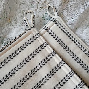 May include: Two square off-white pot holders with black leaf-like patterns and hanging loops. The pot holders have a quilted appearance and are made of a textured fabric. The background is a white lace fabric.