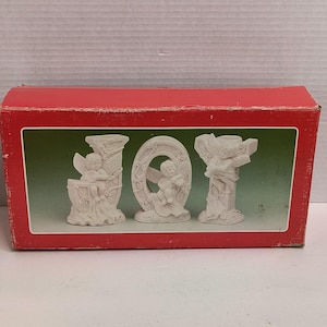 May include: A red box with a green interior houses three white ceramic figurines spelling "JOY". Each figurine depicts an angel. The box is on a white surface.