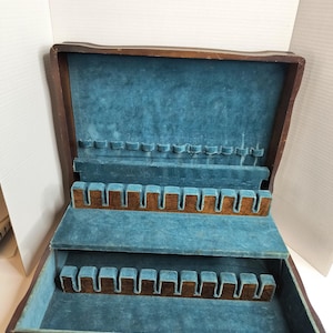 May include: A vintage wooden silverware chest with a dark brown exterior and a teal velvet interior. The chest is open, revealing multiple compartments designed to hold various pieces of flatware. The interior features slots and dividers for organized storage.