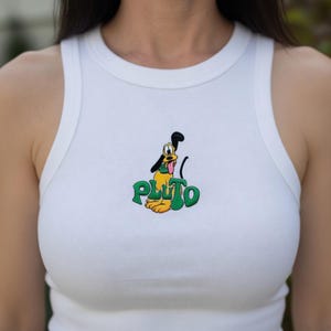 May include: White ribbed tank top featuring a colorful embroidered Pluto graphic. The graphic includes Pluto's image and the word "PLUTO" in green. The tank top is sleeveless and has a round neckline.