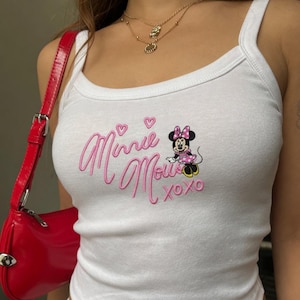 Peut inclure: Débardeur blanc avec un motif Minnie Mouse. Le motif présente les mots "Minnie Mouse" en cursive rose, ainsi qu'une image de dessin animé de Minnie Mouse et le texte "xoxo". Un sac à main rouge est visible dans l'image.