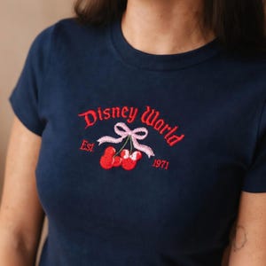 May include: Navy blue t-shirt with embroidered "Disney World" logo in red script, featuring a pink bow and red cherries. The text "Est. 1971" is also embroidered in red. The shirt has short sleeves and a crew neck.