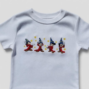 May include: White short-sleeved t-shirt with an embroidered design of four cartoon characters in red robes and blue wizard hats. The design includes yellow stars. The t-shirt is made of a soft, comfortable material.