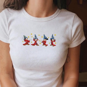 May include: White t-shirt with an embroidered design of Mickey Mouse as a sorcerer. The design shows four Mickey Mouse figures in red robes and blue wizard hats, with yellow star accents.