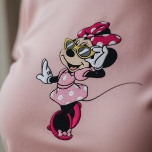 May include: Pink fabric featuring an embroidered Minnie Mouse design. Minnie is wearing sunglasses, a pink dress with white polka dots, and a matching bow. The design is detailed with black, white, and yellow accents.