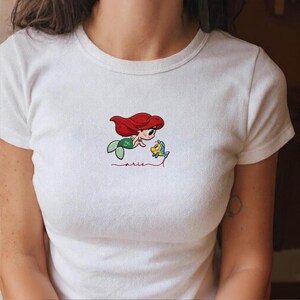 May include: White t-shirt with an embroidered design of a cartoon mermaid with red hair and a green tail, alongside a yellow fish. The word "Ariel" is embroidered in red script beneath the characters.