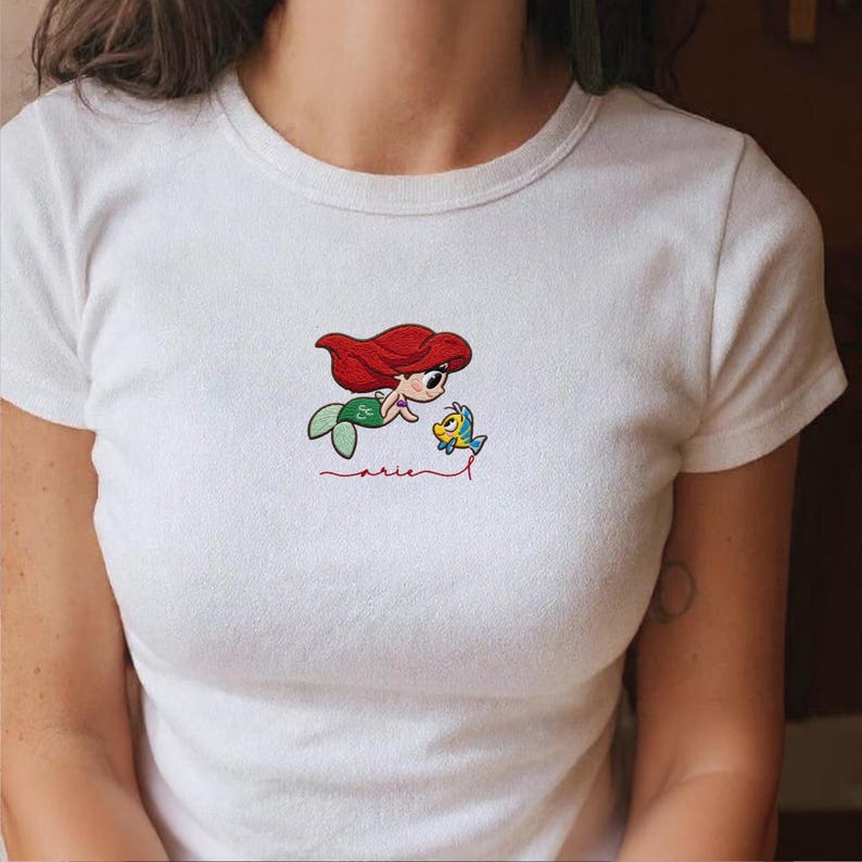 May include: White t-shirt with an embroidered design of a cartoon mermaid with red hair and a green tail, alongside a yellow fish. The word "Ariel" is embroidered in red script beneath the characters.