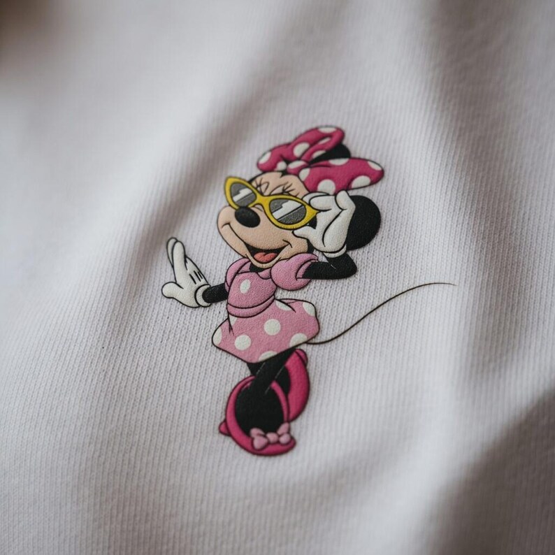 May include: A white sweatshirt featuring a Minnie Mouse graphic. Minnie is depicted in a pink polka-dot dress, matching bow, and yellow sunglasses. The cartoon character is in a pose with one hand raised and the other touching her sunglasses.