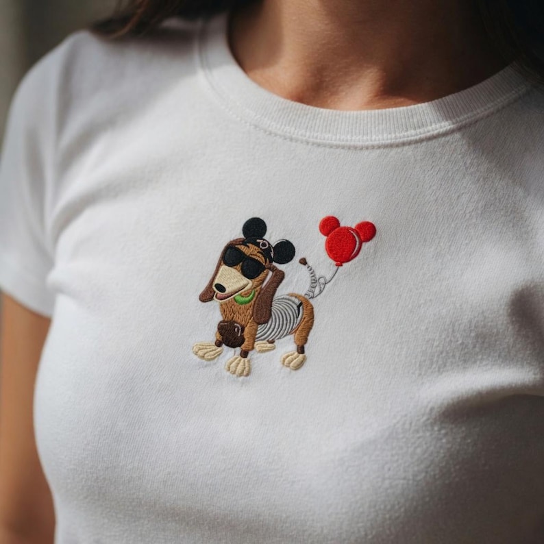 May include: White t-shirt featuring an embroidered cartoon dog wearing sunglasses and Mickey Mouse ears, holding a red balloon. The dog has brown fur, a green collar, and a striped body. The design is centered on the chest.