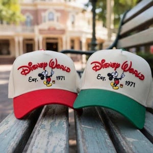 Op de afbeelding: Twee baseballcaps met de woorden "Disney World" in rode letters, het beeld van Mickey Mouse en het jaar "1971". Eén cap heeft een rode klep, de andere een groene klep. Beide caps zijn crèmekleurig.