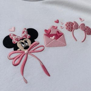 May include: Embroidered design on a white fabric featuring Minnie Mouse with a pink bow, a pink envelope with hearts, and a pink polka dot headband with a bow. The design is in shades of pink and black.