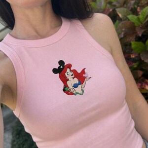 May include: A light pink ribbed tank top featuring a cartoon illustration of Ariel from The Little Mermaid wearing a Mickey Mouse ear headband. The illustration includes a blue seashell bra, red hair, and a green frog.