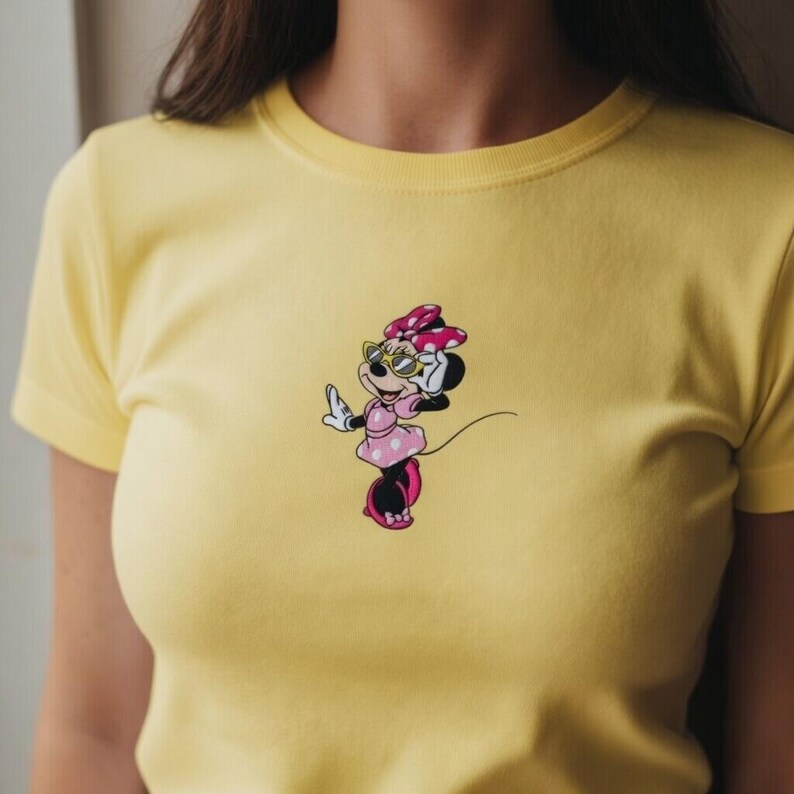 May include: A yellow t-shirt featuring a cartoon Minnie Mouse design. Minnie is depicted wearing sunglasses, a pink dress with white polka dots, and a matching bow. The design is centered on the front of the shirt.