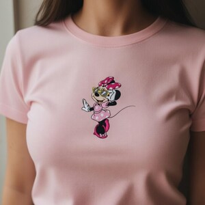 May include: A light pink t-shirt featuring a cartoon Minnie Mouse design. Minnie Mouse is wearing sunglasses, a pink dress with white polka dots, and a matching bow. The shirt has short sleeves and a crew neck.
