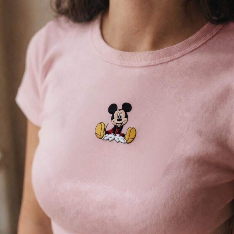 K&ouml;nnte beinhalten: Hellrosa T-Shirt mit einem kleinen, gestickten Bild von Mickey Mouse. Die Zeichentrickfigur ist in sitzender Position dargestellt und tr&auml;gt rote Shorts, gelbe Schuhe und wei&szlig;e Handschuhe. Das Shirt hat einen Rundhalsausschnitt und kurze &Auml;rmel.
