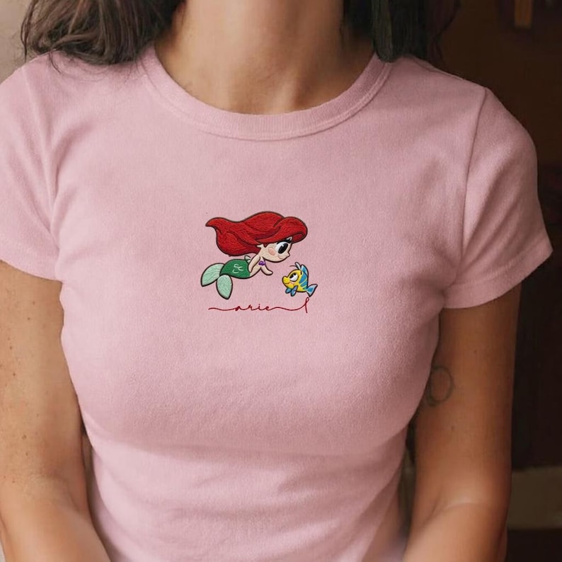 May include: A pale pink t-shirt with an embroidered design. It depicts a cartoon mermaid with red hair and a green tail, alongside a yellow fish. The word "Ariel" is written in cursive below the characters.